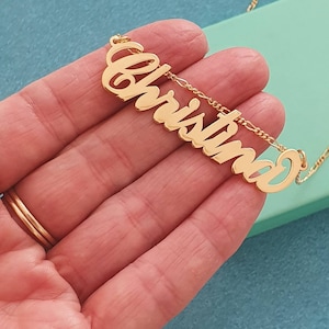May include: A gold-colored necklace with the name "Christina" in a cursive font. The name is the focal point, suspended from a delicate chain. The necklace is displayed against a light green and blue background.