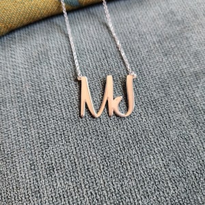 Peut inclure: Un collier en argent avec les initiales "MJ" en écriture cursive. Le pendentif, de couleur or clair, est suspendu à une fine chaîne en argent. Le collier est présenté sur un fond gris texturé.