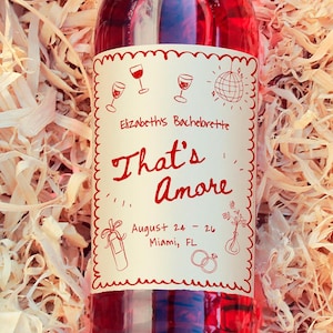 Puede incluir: Una botella de vino tinto con una etiqueta decorativa que dice "That's Amore" en una caja de madera llena de virutas de madera. La etiqueta también incluye el texto "Elizabeths Bachelorette" y la fecha "24-26 de agosto Miami, FL."