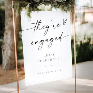 May include: White sign with black calligraphy that reads "They're engaged" with a heart symbol. Below the text is "Let's Celebrate!" and "Nicole & Eric". The sign is on a copper stand.
