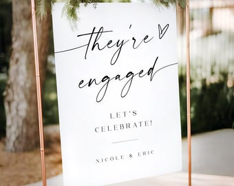 May include: White sign with black calligraphy that reads "They're engaged" with a heart symbol. Below the text is "Let's Celebrate!" and "Nicole & Eric". The sign is on a copper stand.
