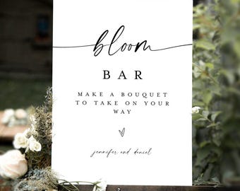 May include: A white sign on a wooden easel with the words "bloom BAR" in black script. Below, it reads "MAKE A BOUQUET TO TAKE ON YOUR WAY" with a heart symbol and names "jennifer and daniel". Soft focus background.