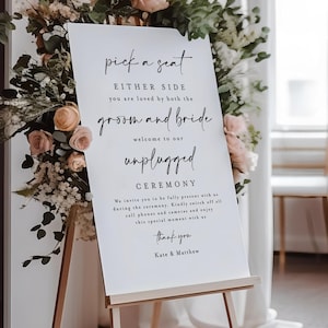 Puede incluir: Un letrero de ceremonia de boda sobre un caballete de madera. El letrero dice: "Elige un asiento, a ambos lados, eres amado por los novios, ceremonia desconectada". Arreglos florales con flores rosas y blancas rodean el letrero.