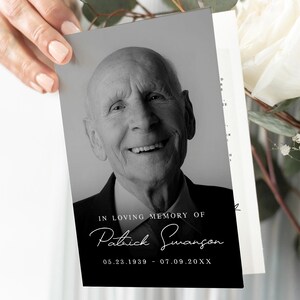 May include: A black and white memorial card with a portrait of an elderly man. The card includes the text "IN LOVING MEMORY OF Patrick Swanson 05.23.1939 - 07.09.20XX". The card is held near a bouquet of white flowers and foliage.