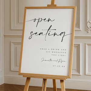 Op de afbeelding: Houten ezel met een wit bord met de tekst "open seating" in elegant zwart schrift. Daaronder staat "Grab a drink and sit wherever you like!" gevolgd door namen en een datum. Het bord is omlijst in licht hout.