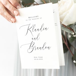 Puede incluir: Dos tarjetas de bienvenida de boda blancas con texto de caligrafía negra que dice "Bienvenida a la boda de Klaudia y Brandon", además de la ubicación y la fecha. Las tarjetas se sostienen frente a un ramo de rosas blancas y vegetación.