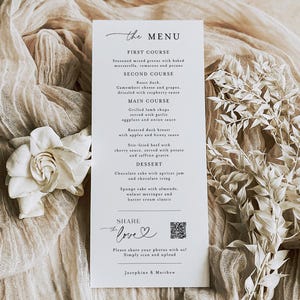 May include: A white wedding menu card with a refined script font, listing a detailed menu. The menu includes dishes such as roast duck and sponge cake. The card is decorated with a white flower and sprigs, set against a neutral fabric backdrop.