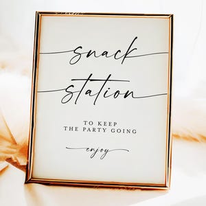 May include: A rectangular sign with a gold frame, displaying the words "Snack Station" in black cursive. Beneath, it reads "To keep the party going" and "enjoy". The sign is set against a soft, white backdrop.