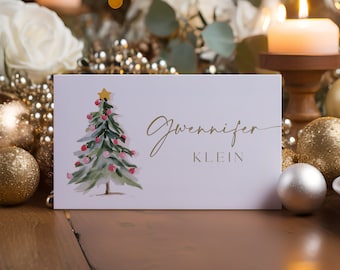 Puede incluir: Una tarjeta de lugar blanca con letras doradas que dice "Gennifer Klein". La tarjeta presenta una pintura de acuarela de un árbol de Navidad con adornos rosados y una estrella dorada en la parte superior. La tarjeta está rodeada de decoraciones navideñas doradas y blancas.