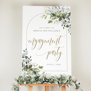 May include: A white engagement party sign with gold lettering and a floral design. The sign reads "Welcome to Amelia and William's engagement party" with the date June 6, 20XX. The sign is on a wooden easel and decorated with greenery.
