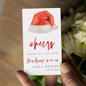 May include: A white card with a watercolor image of a red Santa hat and the word "cheers" in red script. The card reads "Redeem for 1 free drink" and "This drink is on us" with the names "Kate & Matthew" and the date "12.23.XX".