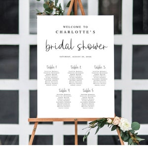 May include: A white sign with black text announcing a bridal shower for Charlotte on August 20th. The sign lists the names of guests assigned to each table.