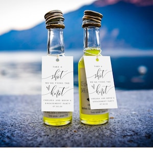May include: Two small glass bottles with white labels that say "Take a shot, we're tying the knot" with a heart and the text "Chelsea and Kevin's Engagement Party" and the date "07.23.23". The bottles are filled with a yellow liquid and are sitting on a brown surface.