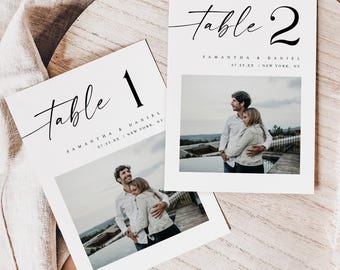 May include: Two white wedding table cards with black text. The cards read "Table 1" and "Table 2" in a script font. Each card has a photo of a couple. The cards also include the names "Samantha & Daniel" and the date and location.