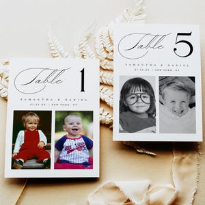 May include: Two white table number cards with photos of children. The cards read "Table 1" and "Table 5" in elegant script. The cards also include the names "Samantha & Daniel" and a date and location. The background is a neutral beige.