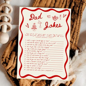 May include: A cream-colored card with red text that reads "Dad Jokes" with a list of jokes. The card is surrounded by a red border and rests on a brown onesie with a wooden rattle.