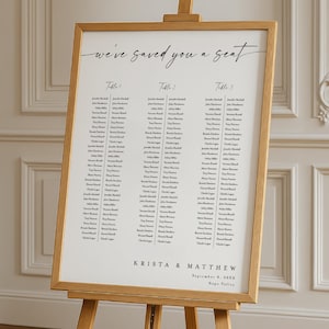 May include: A wooden easel displaying a seating chart with the text "we've saved you a seat." The chart lists names under the headings "Table 1," "Table 2," and "Table 3." The names are in black font. The names "Krista & Matthew" and the date are at the bottom.