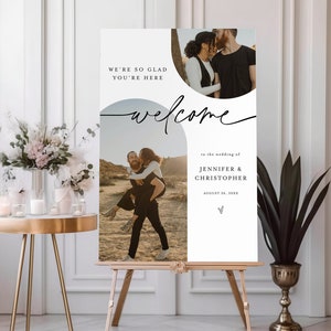 May include: A white welcome sign with a black script font for a wedding. The sign features two photos of a couple in a desert setting. The text on the sign reads "We're so glad you're here, welcome to the wedding of Jennifer & Christopher, August 26, 20XX".