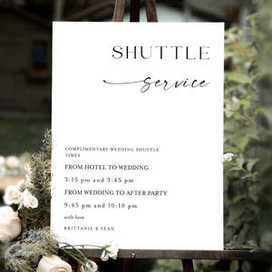 May include: A white sign on a wooden easel with the words "SHUTTLE SERVICE" in black. The sign provides shuttle times for a wedding, including times from the hotel to the wedding and from the wedding to the after-party. Floral arrangements decorate the base.