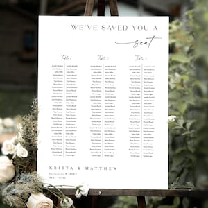 Puede incluir: Un plano de asientos de boda blanco sobre un caballete, con el texto "WE'VE SAVED YOU A seat". El gráfico enumera los nombres bajo los encabezados "Table 1", "Table 2" y "Table 3". Los nombres están en fuente negra. Los nombres Krista & Matthew están en la parte inferior.