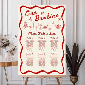 May include: A cream-colored wedding seating chart with red decorative border and text. The words "Ciao Bambino" are at the top, with "Please Take a Seat" below. Tables 1-6 are listed with names. The chart is on an easel.