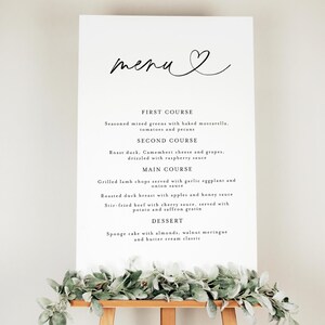 May include: A white menu sign with the word "menu" in cursive script and a heart symbol. The menu lists a first course, second course, main course, and dessert. The sign is on a wooden easel, decorated with greenery.