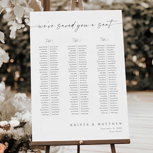 Op de afbeelding: Een witte zitplaatsindeling op een houten ezel met de tekst "we've saved you a seat". De tabel bevat namen onder de titels "Tafel 1", "Tafel 2" en "Tafel 3". De namen zijn voor Krista & Matthew, 8 september 20XX, Napa Valley.
