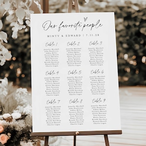 Puede incluir: Un plano de asientos de boda blanco sobre un caballete de madera. El gráfico presenta una elegante caligrafía con las palabras "Our favorite people" y los nombres de los invitados organizados por número de mesa. Los nombres están en una fuente sencilla y legible.