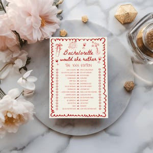 May include: A white card with red text and a decorative border. The card reads "Bachelorette would she rather THE XXX EDITION" with a list of playful questions. The card is surrounded by flowers and decorative items.