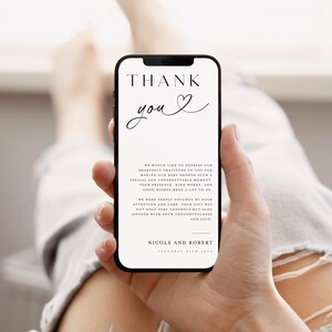 May include: A white thank you card with black text and a heart symbol. The text reads "Thank you" and includes a message expressing gratitude for attending a baby shower. The card is held in a hand, with a portion of a person's leg visible in the background.