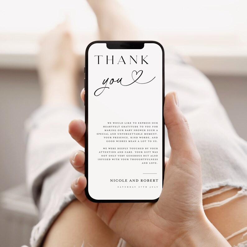 May include: A white thank you card with black text and a heart symbol. The text reads "Thank you" and includes a message expressing gratitude for attending a baby shower. The card is held in a hand, with a portion of a person's leg visible in the background.