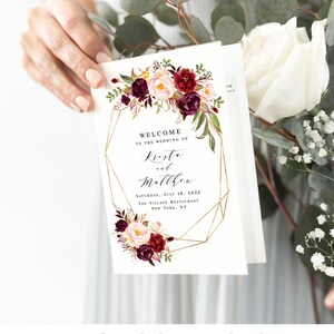 May include: Wedding program with a gold geometric frame and burgundy and blush floral design. The text reads "Welcome to the wedding of Krista and Matthew, Saturday, July 16, 2022, The Village Restaurant, New York, NY."