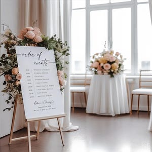 Puede incluir: Un caballete blanco que muestra un letrero de orden de eventos de boda, adornado con arreglos florales. El letrero presenta una caligraf&iacute;a elegante y un horario, que incluye la ceremonia, fotos y detalles de la recepci&oacute;n. El letrero est&aacute; en un espacio luminoso y espacioso con grandes ventanas.