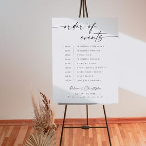 Puede incluir: Un letrero blanco de evento de boda sobre un caballete negro. El letrero muestra las palabras "order of events" en una escritura elegante, seguido de un programa detallado que incluye la ceremonia de boda, la cena y el baile. Tambi&eacute;n aparecen los nombres "Rebecca & Christopher".
