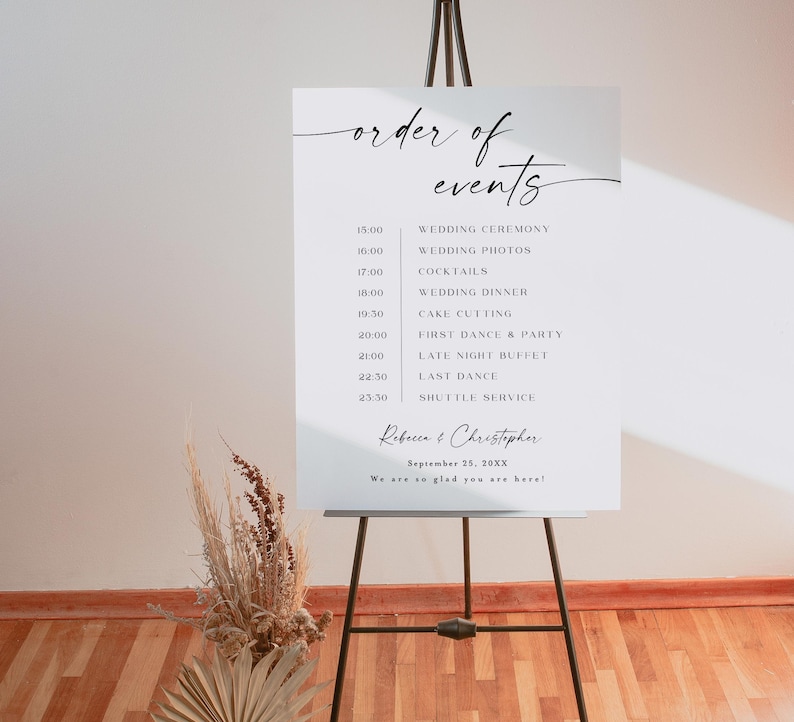 Puede incluir: Un letrero blanco de evento de boda sobre un caballete negro. El letrero muestra las palabras "order of events" en una escritura elegante, seguido de un programa detallado que incluye la ceremonia de boda, la cena y el baile. Tambi&eacute;n aparecen los nombres "Rebecca & Christopher".