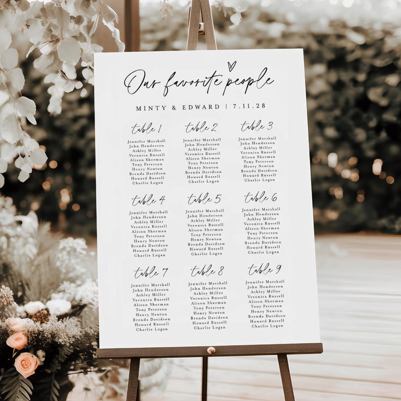 Stylish outdoor wedding seating chart displayed on a dark wood easel among blooming floral arrangements. The design features a clean layout for up to nine tables, customized with names and wedding date, ideal for modern receptions.