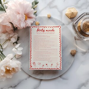Op de afbeelding: Een cr&egrave;mekleurig kaartspel getiteld "Dirty Minds" met rode tekst en een decoratieve rand. Het spel is omgeven door roze en witte bloemen, gouden geometrische vormen en een glazen pot op een marmeren oppervlak.