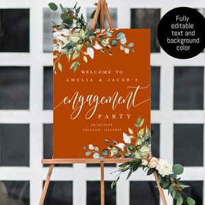 Puede incluir: Un cartel de bienvenida para una fiesta de compromiso con un fondo naranja &oacute;xido y texto blanco. El texto dice "Bienvenido a Amelia & Jacob's Engagement Party 08.20.20XX Chicago, Illinois". El cartel est&aacute; decorado con vegetaci&oacute;n acuarela.