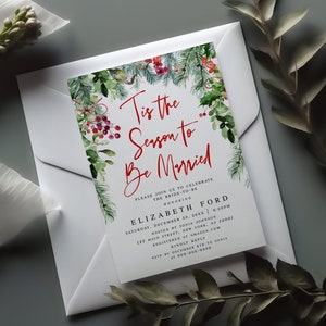May include: A white invitation card with the text "Tis the Season to Be Married" in red script, framed by watercolor greenery and red berries. The card is inside a white envelope, perfect for a winter wedding.