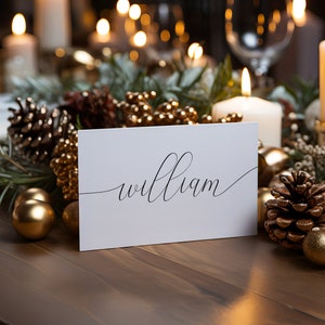 Puede incluir: Una tarjeta blanca con el nombre "william" escrito en cursiva. La tarjeta est&aacute; rodeada de ramas de pino, pi&ntilde;as y adornos dorados.