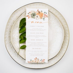 Op de afbeelding: Een printbare Thanksgiving-menukaart met een aquarel-illustratie van pompoenen en herfstbladeren. Het menu bevat een voorgerecht, een hoofdgerecht, bijgerechten en een dessert.