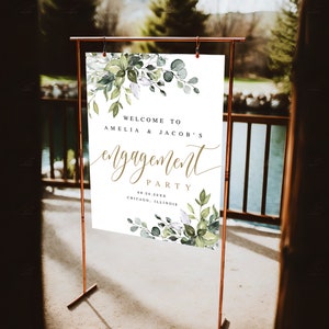 Puede incluir: Un cartel de bienvenida blanco con follaje verde acuarela y letras doradas. El cartel dice "Bienvenido a Amelia & Jacob's Engagement Party 08.20.20XX Chicago, Illinois".