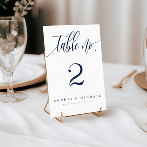 May include: White table number card with a gold stand. The card has a navy blue cursive font that says "Table No. 2" and "Sophia & Michael, Saturday 27th July".