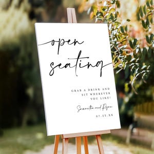 May include: A white sign on a wooden easel with the words "open seating" in elegant black script. Below, it reads "Grab a drink and sit wherever you like!" followed by names and a date.