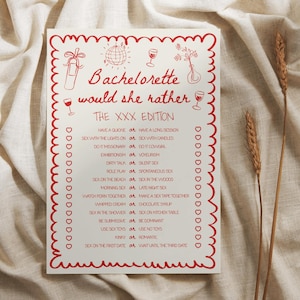 May include: A white card with red border and text that reads "Bachelorette would she rather THE XXX EDITION". The card has red illustrations of a disco ball, wine glasses, and flowers. The card is on a beige fabric background.