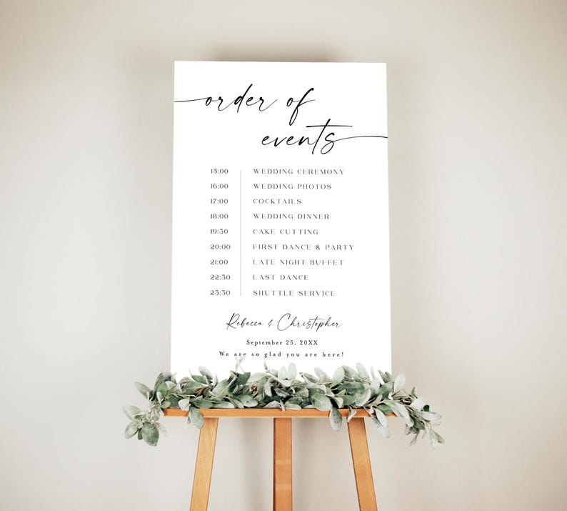 Puede incluir: Un letrero de boda blanco con las palabras "order of events" en una elegante escritura. El letrero enumera el programa de la boda, incluyendo la ceremonia, fotos, cena y baile. El letrero est&aacute; sobre un caballete de madera, decorado con vegetaci&oacute;n.
