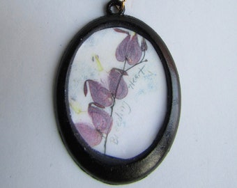 May include: A black oval pendant necklace with a black cord. The pendant features a pressed flower design of a bleeding heart flower with the text "Bleeding Heart" written on the paper.