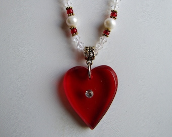 May include: A red heart-shaped pendant necklace with a clear crystal in the center. The necklace is made with clear and red beads and white pearls.
