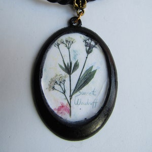 Op de afbeelding: Een zwarte koordketting met een ronde zwarte hanger met een geperst bloemenontwerp en de tekst "sweet Woodruff".