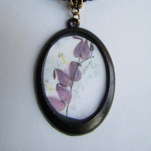 May include: A black oval pendant necklace with a black cord. The pendant features a pressed flower design of a bleeding heart flower with the text "Bleeding Heart" written on the paper.